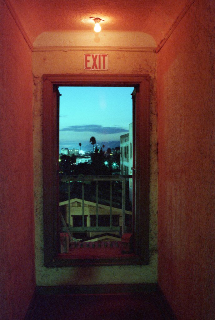 A fire escape doorway, lit by a single bulb. Through the open door can be seen Hollywood at dusk, with buildings, streetlights and palm trees. 