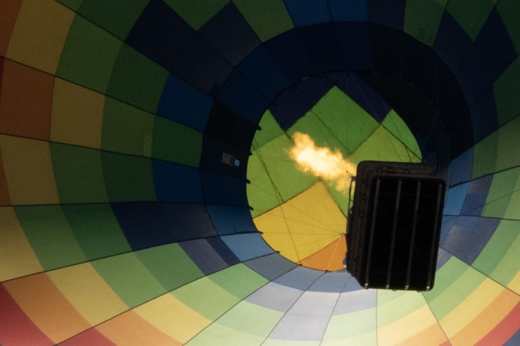 From under the gondola of a hot air balloon in flight a plume of yellow flame reaches up.