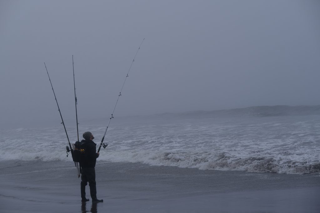 A lone fisherman stands on the beach looking up, while the surf and sky blend together in the fog.