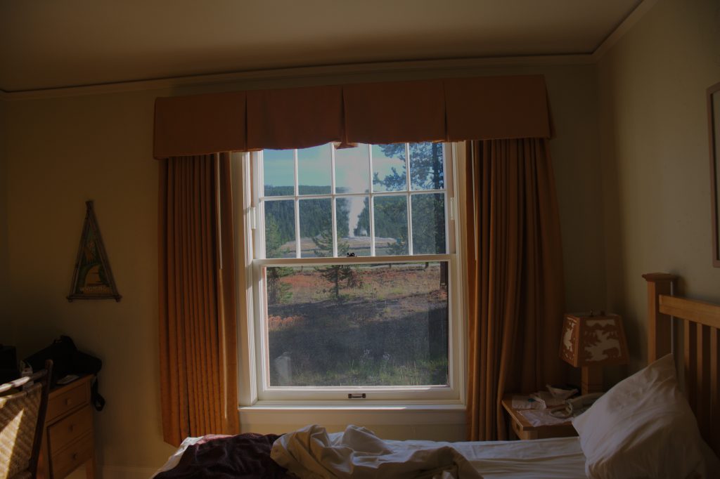 Old Faithful geyser erupts outside the dimly lit hotel room, framed by the screen window.