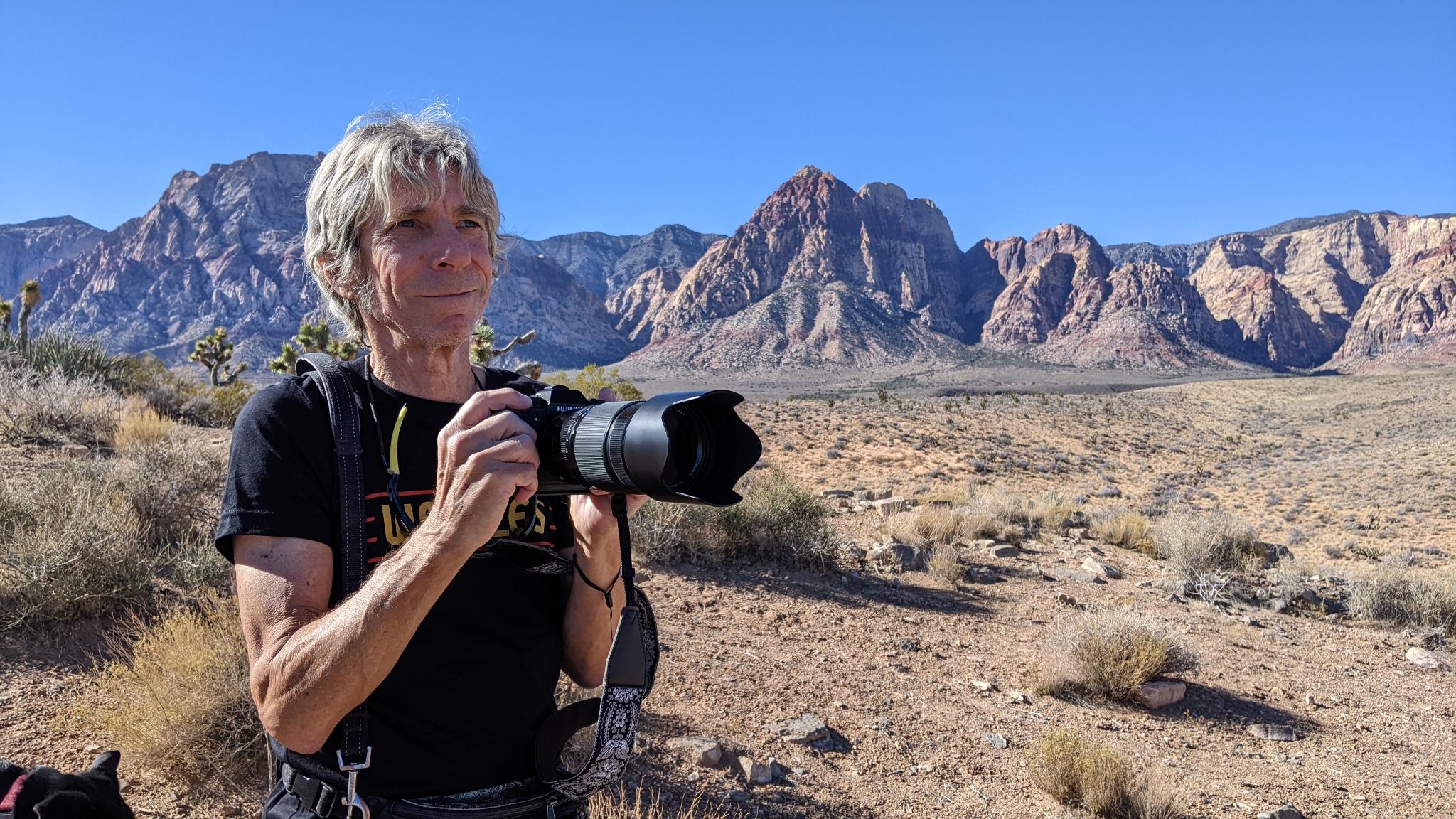 Artist holds camera and looks off into the distance in a desert scene with rocky outcroppings in the background.