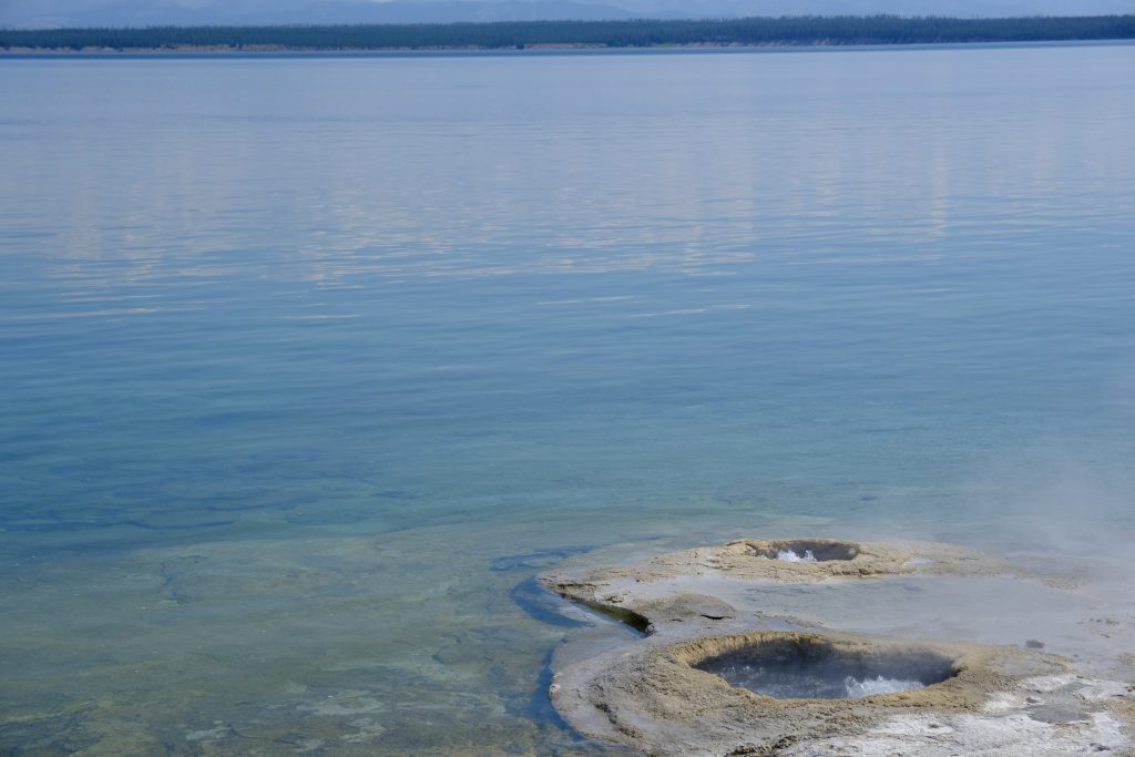 Two crater-like openings in the rock at the edge of an azure lake. Inside the craters, water boils. Vapor drifts off. At the far side of the lake, forests of pine, and a sliver of sky.