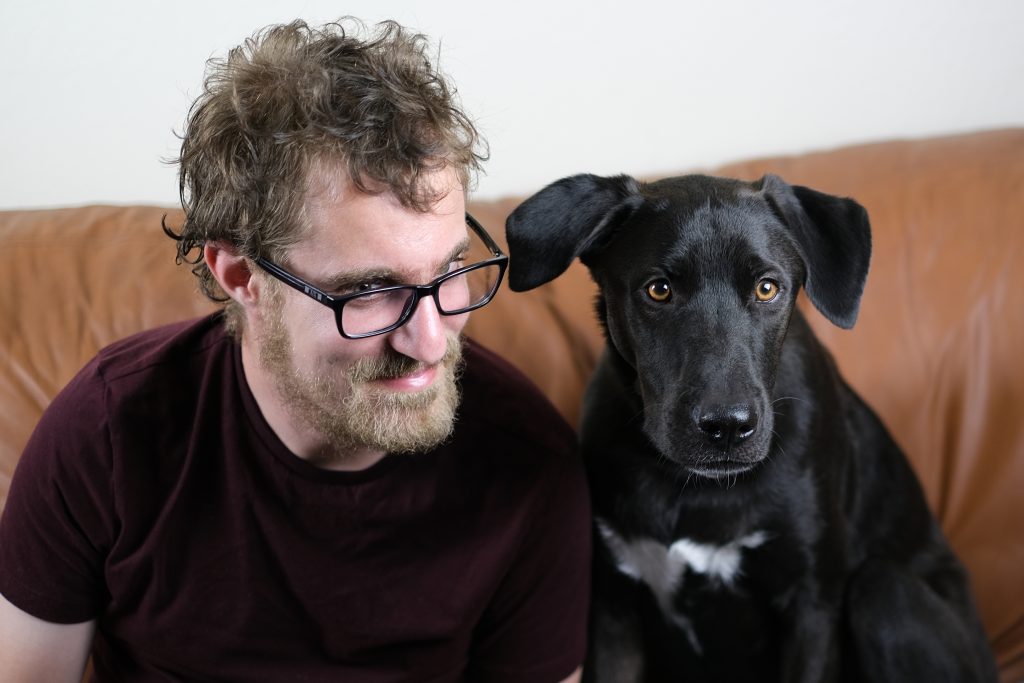 A bearded young man with glasses wears a pleased expression while sitting next to his black dog with brown eyes and a white design on its chest.