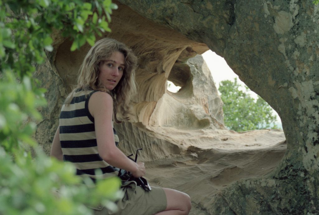 Attractive woman in her 20's looks back over her right shoulder while framed by vegetation on the left and a rock wall on the right.  Between her and the rock wall is an opening to the light.