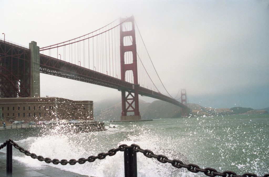 A large metal chain link is supported by a couple of metal posts, while the surf crashes behind. The water of the bay is green. A two story large brick building with many window openings sits at the foot of the Golden Gate Bridge. The sky is a low fog, which partially obscures the highest parts of the two bridge towers.  On the far side, a hint of blue sky.