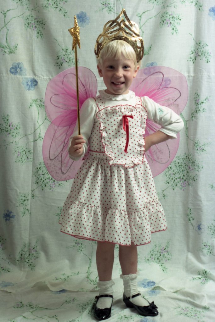 A young girl looks extremely happy to be wearing a gold crown and pink fairy wings, while holding a magic wand with a star on top.