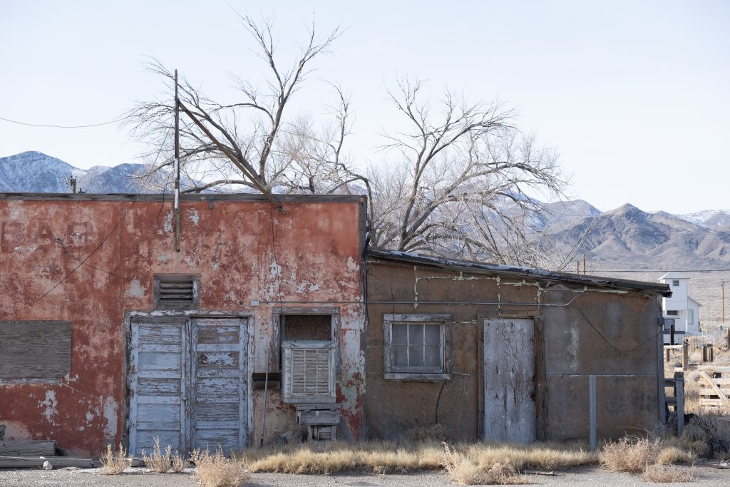 An old air conditioner hangs low in the partially boarded up window. Weeds grow up from the driveway in front of three doors. The two adjoining doors have peeling grey paint, while the third appears weathered and unpainted. The main building has faded red paint, while the building addition may have been painted brown or maybe was never painted. Behind the building is a bare tree, and beyond are dense hills, the tallest of which are lightly covered in snow. 