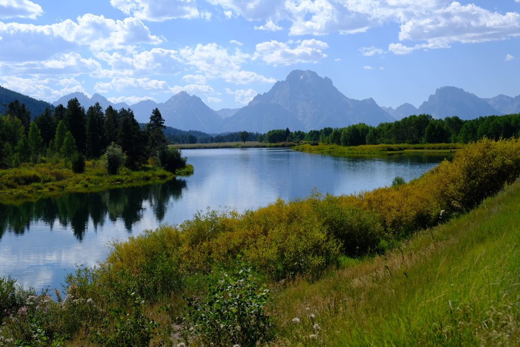 A green hill slopes to a winding river with pine trees on the other side. In the background are majestic rock mountains, and the cloudy sky is reflected in the water.