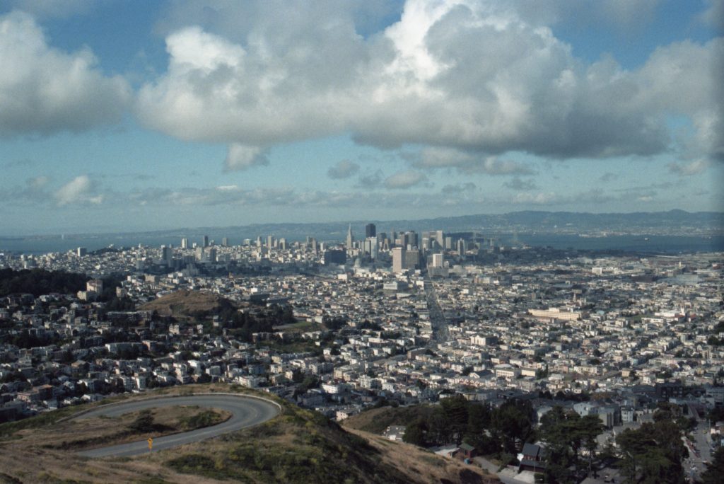 View from a large hill in San Francisco toward downtown and the East Bay. Part of the winding road to the top is visible, as are many individual buildings and the Bay Bridge. The sky is blue with large clouds.