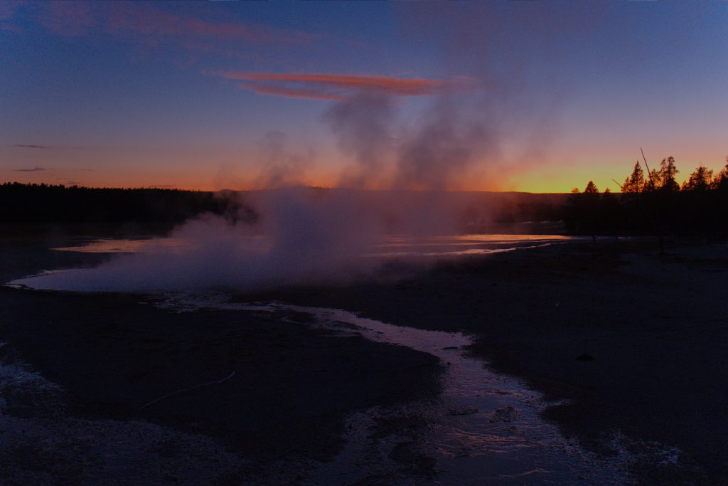 The sun has already set, but a little light remains. In the shy some wispy clouds appear reddish, as does most of the vapor cloud coming from a volcanic feature nearby.  There is a reflection of the sky in water behind the vapor cloud, as well as in a stream heading towards us.