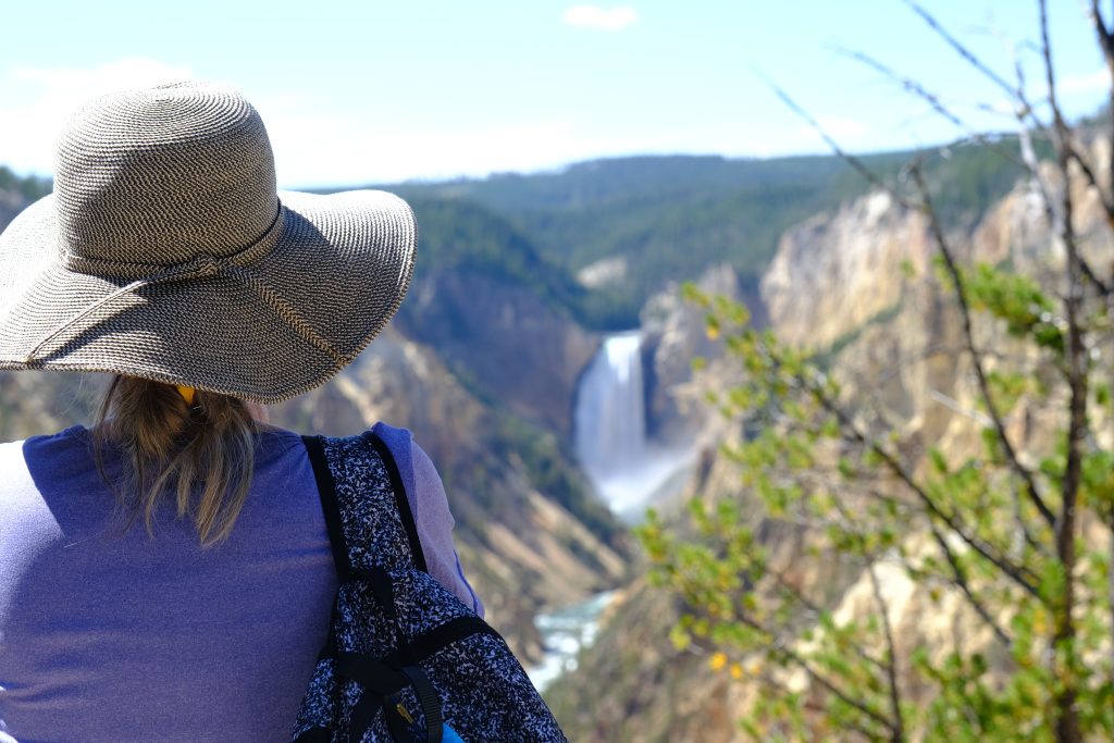 A woman wearing a large sun hat gazes at a large waterfall in the distance.