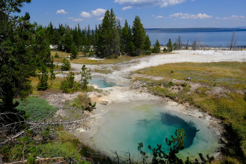 We are near an emerald green pond.  To the left and ahead are pine trees and a smaller green pond.  The sky is light blue with small clouds, and off in the distance on the right we get a peek of a blue lake with low hills beyond.