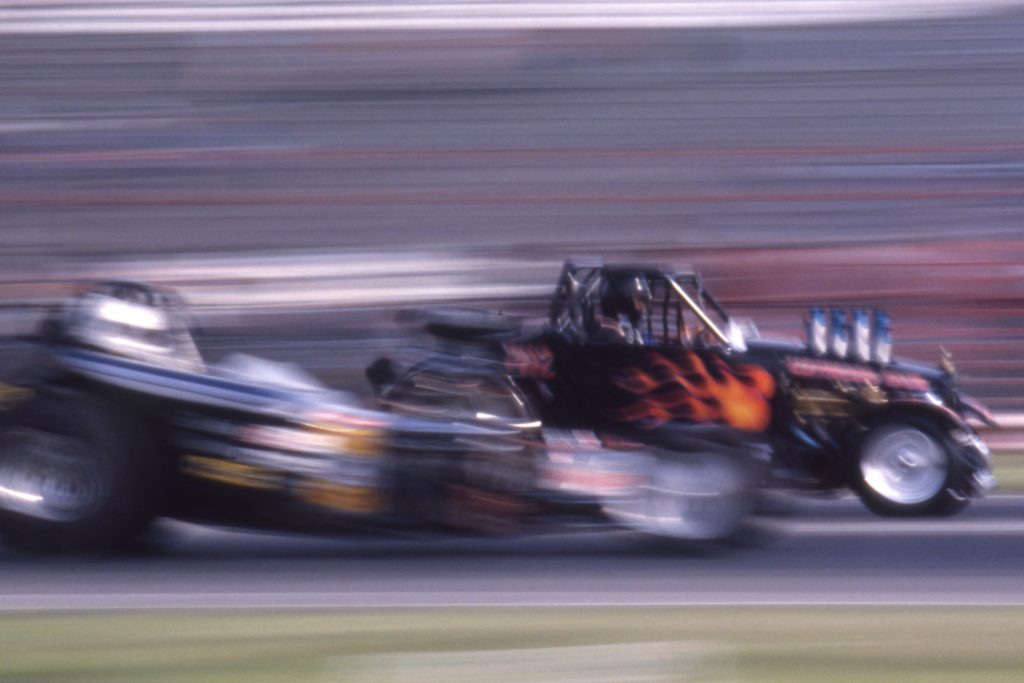 Two hotrods race on a track, with the background blurred by the camera panning with the cars.