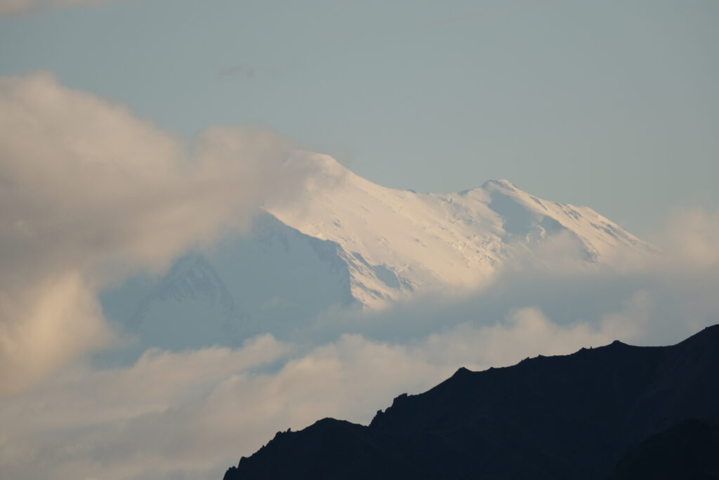 Snow covered Denali peak rises above nearby hills, partially obscured by cloud