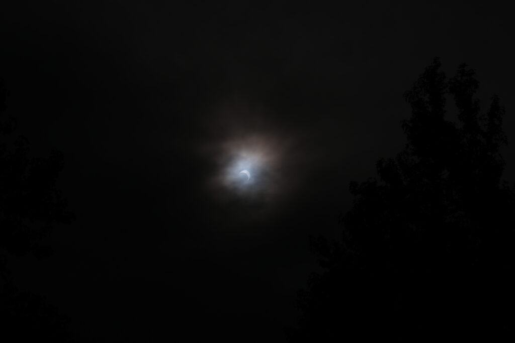 Partial eclipse of the sun, peaking through clouds with dark trees silhouetted on both sides 